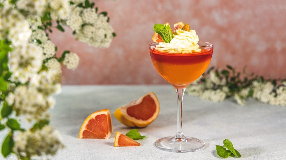 Glas mit orangem Wackelpudding und Sahne, dekoriert mit Minze und Zitrus, auf Betonfläche mit weißen Frühlingsblüten.