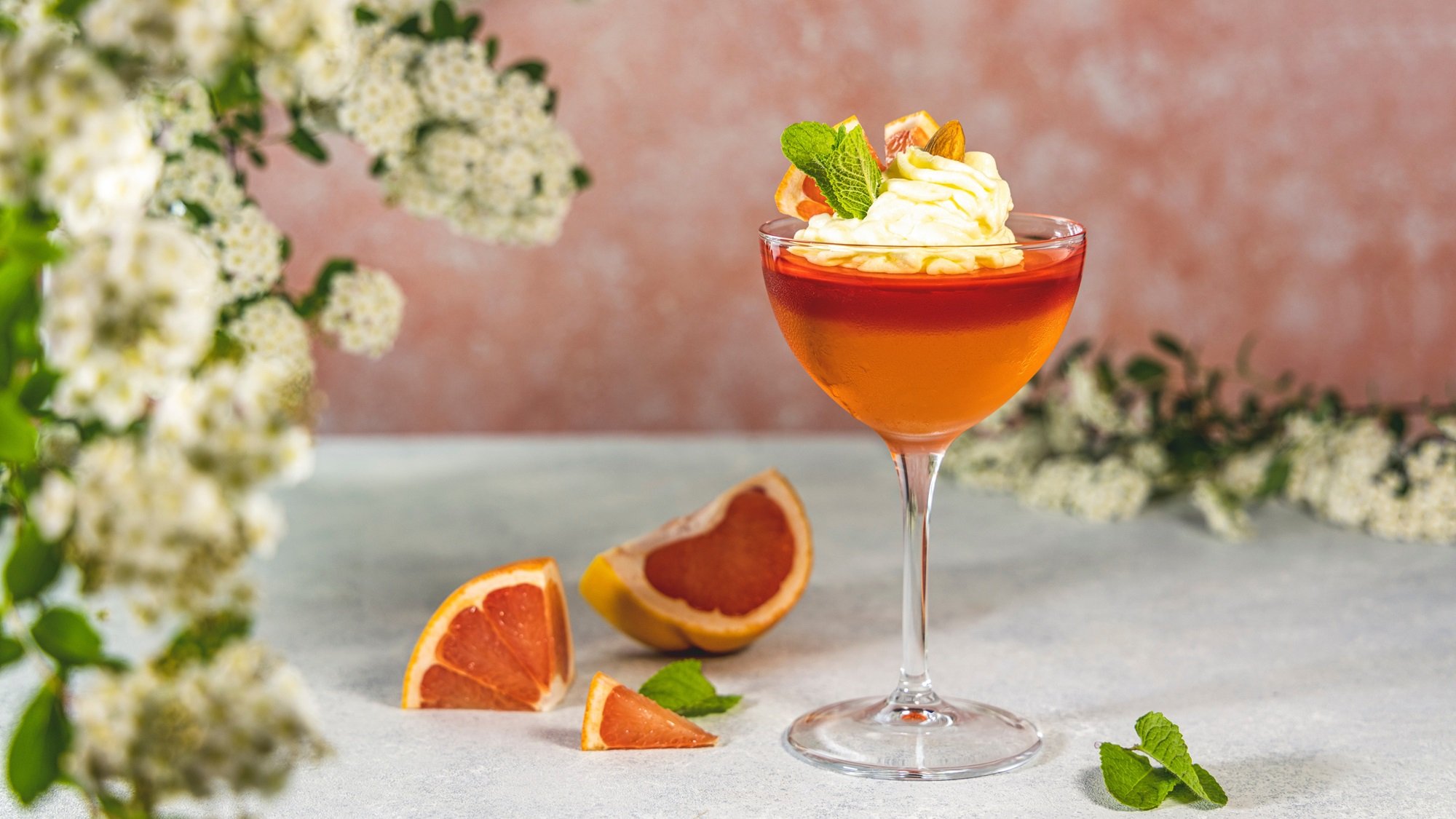 Glas mit orangem Wackelpudding und Sahne, dekoriert mit Minze und Zitrus, auf Betonfläche mit weißen Frühlingsblüten.
