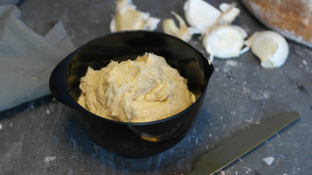 Ein schwarzes Schälchen mit der Knoblauchbutter auf steinernem Untergrund und schwarzem Hintergrund. Alles von schräg oben fotografiert.