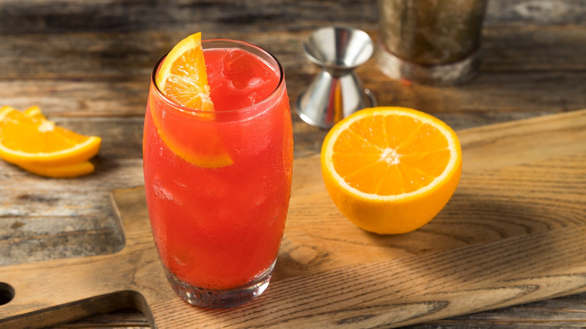 Leuchtender Alabama Slammer mit Orangenscheibe in hohem Glas auf Holzbrett, daneben halbe Orange und Barkeeper-Zubehör.