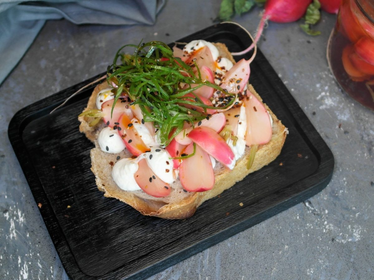 Ein schwarzes Brett mit einer Brotscheibe und dem Radieschensalat als Belag. Alles von schräg oben fotografiert.