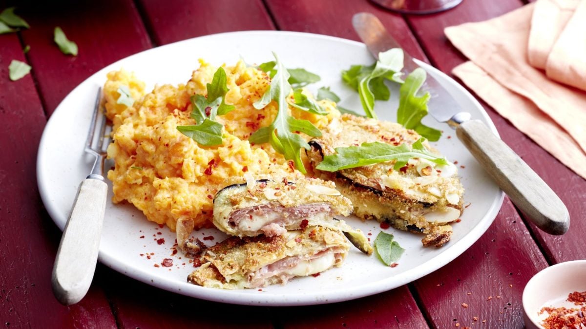 Ein angerichteter Teller mit Auberginen-Cordon Bleu, gefüllt mit Schinken und Käse, dazu ein cremiges Kartoffel-Möhren-Püree und frischer Rucola, serviert auf einem weißen Teller mit Besteck auf einem roten Holztisch.