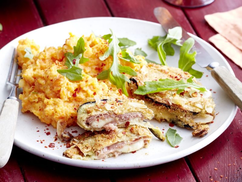 Ein angerichteter Teller mit Auberginen-Cordon Bleu, gefüllt mit Schinken und Käse, dazu ein cremiges Kartoffel-Möhren-Püree und frischer Rucola, serviert auf einem weißen Teller mit Besteck auf einem roten Holztisch.