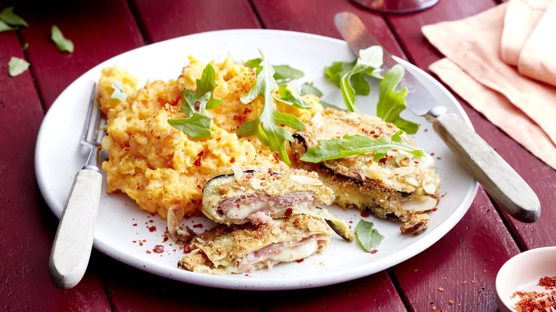 Ein angerichteter Teller mit Auberginen-Cordon Bleu, gefüllt mit Schinken und Käse, dazu ein cremiges Kartoffel-Möhren-Püree und frischer Rucola, serviert auf einem weißen Teller mit Besteck auf einem roten Holztisch.