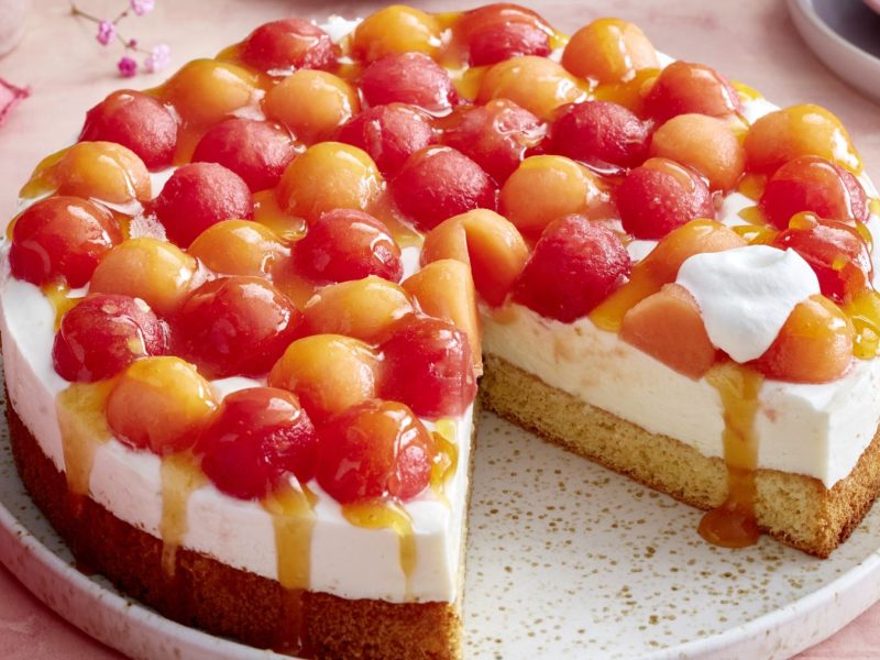 Eine sommerliche Melonen-Joghurt-Torte auf einem Teller, belegt mit roten und orangefarbenen Melonenkugeln, cremiger Joghurtfüllung und lockerem Biskuitboden. Die Torte ist angeschnitten, etwas Fruchtsoße läuft am Rand herab, ein Klecks Sahne liegt auf einem Stück.