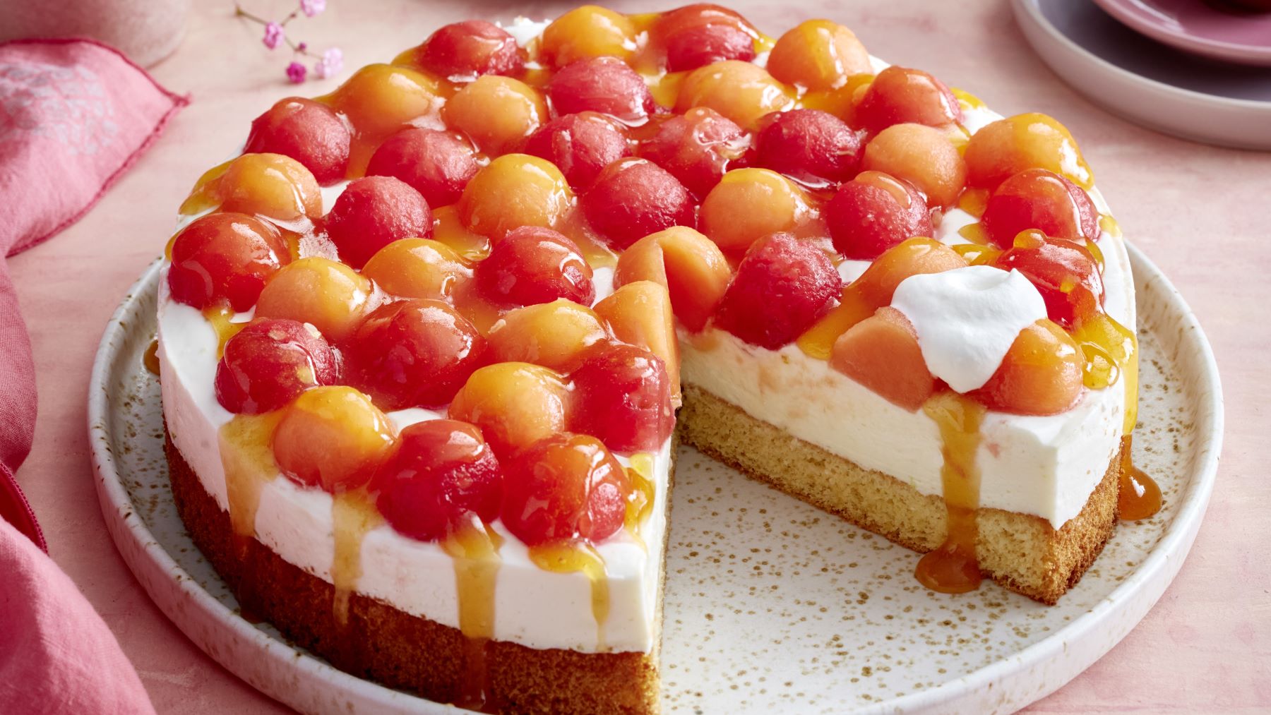 Eine sommerliche Melonen-Joghurt-Torte auf einem Teller, belegt mit roten und orangefarbenen Melonenkugeln, cremiger Joghurtfüllung und lockerem Biskuitboden. Die Torte ist angeschnitten, etwas Fruchtsoße läuft am Rand herab, ein Klecks Sahne liegt auf einem Stück.