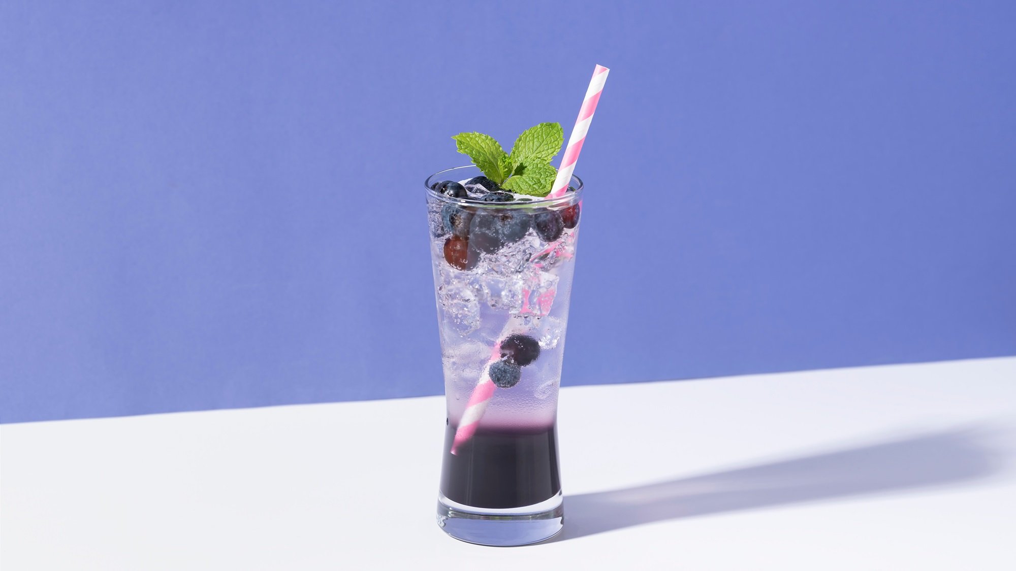 Erfrischender Mocktail mit Blaubeeren, Eis und Minze in einem schlanken Glas auf hellem Untergrund vor blauer Wand.