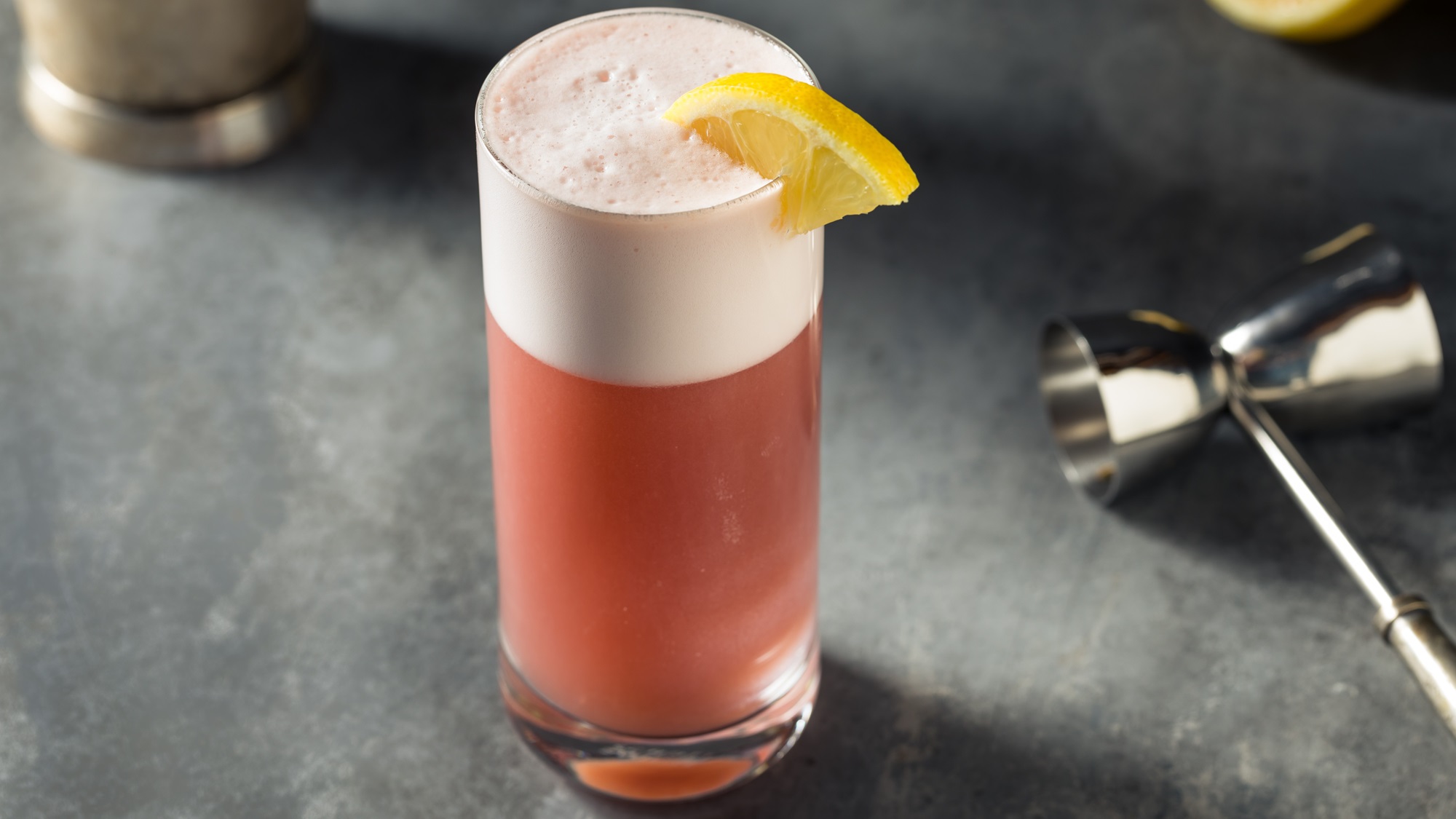 Ein schaumiger Chicago Fizz mit Zitronenschnitz steht auf grauem Steinuntergrund, daneben liegt ein silberner Messbecher.