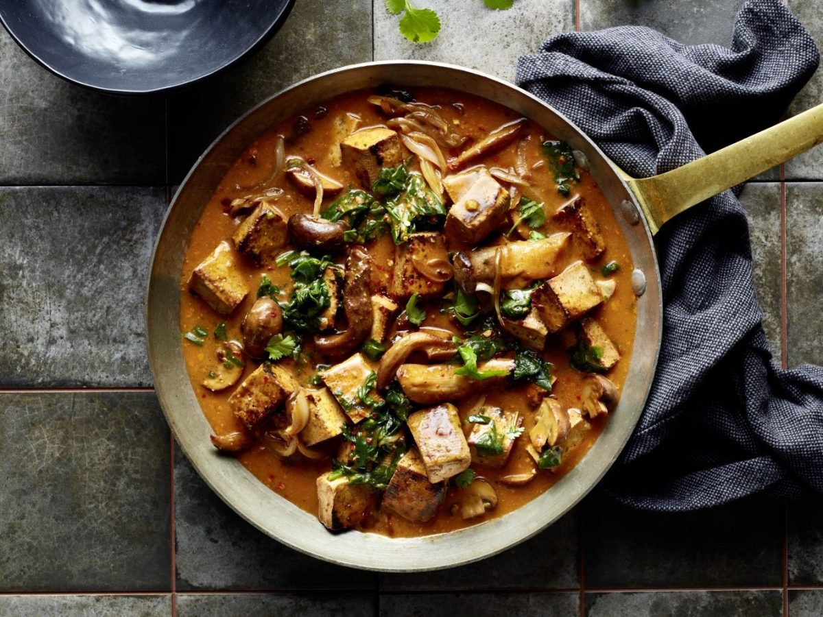 Pfanne mit cremigem Kokos-Pilz-Curry, darin gebratene Tofuwürfel, verschiedene Pilze, Zwiebeln und frische Kräuter auf einem rustikalen Fliesenuntergrund, daneben ein dunkles Tuch.