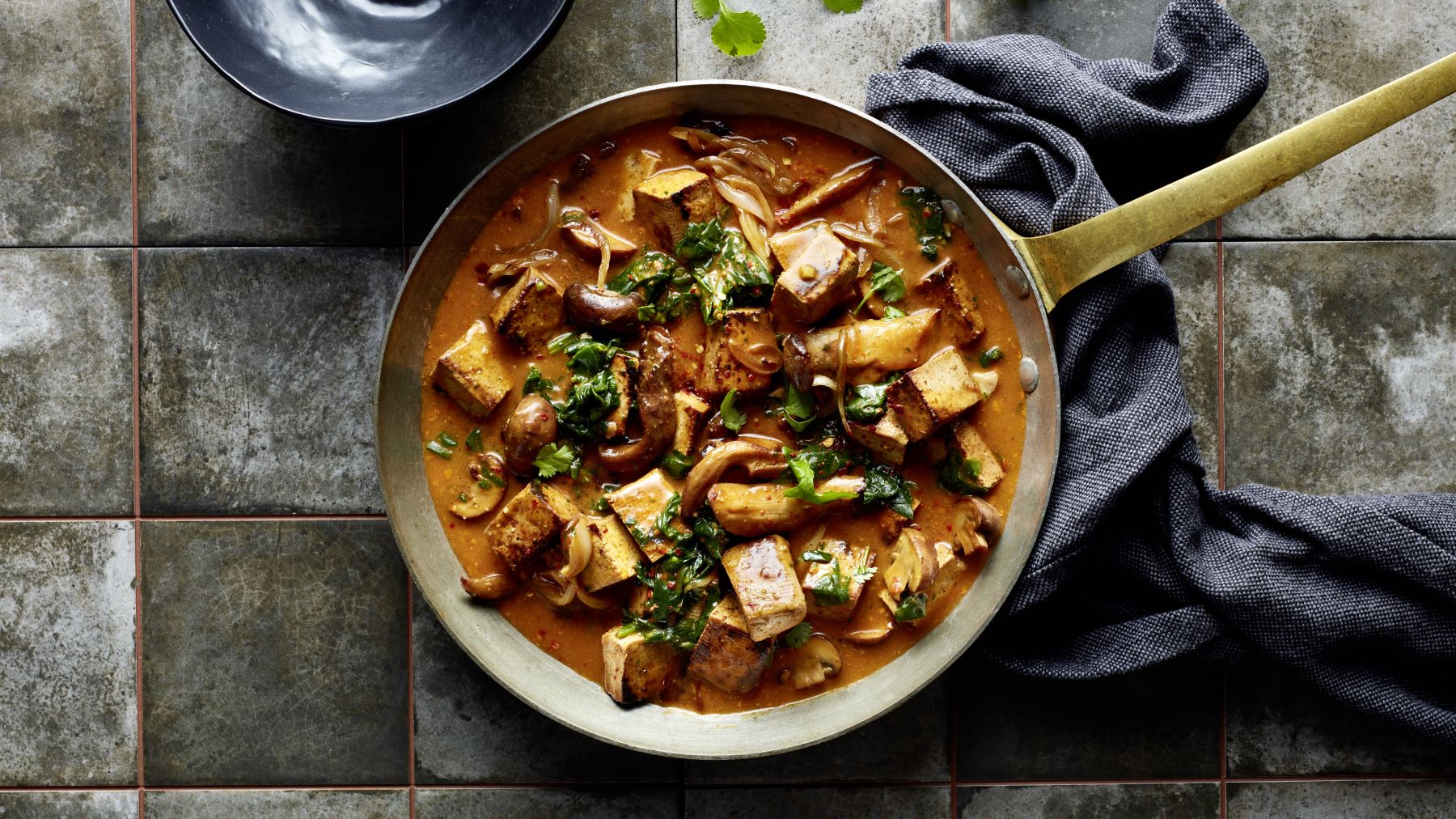 Pfanne mit cremigem Kokos-Pilz-Curry, darin gebratene Tofuwürfel, verschiedene Pilze, Zwiebeln und frische Kräuter auf einem rustikalen Fliesenuntergrund, daneben ein dunkles Tuch.