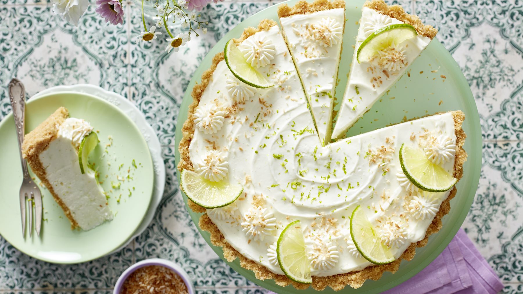 Eine Kokos-Limetten-Tarte mit knusprigem Boden, cremiger Füllung und Sahnetuffs, dekoriert mit Limettenscheiben, gerösteten Kokosraspeln und Limettenschale. Ein Stück ist angeschnitten und liegt auf einem Teller daneben.