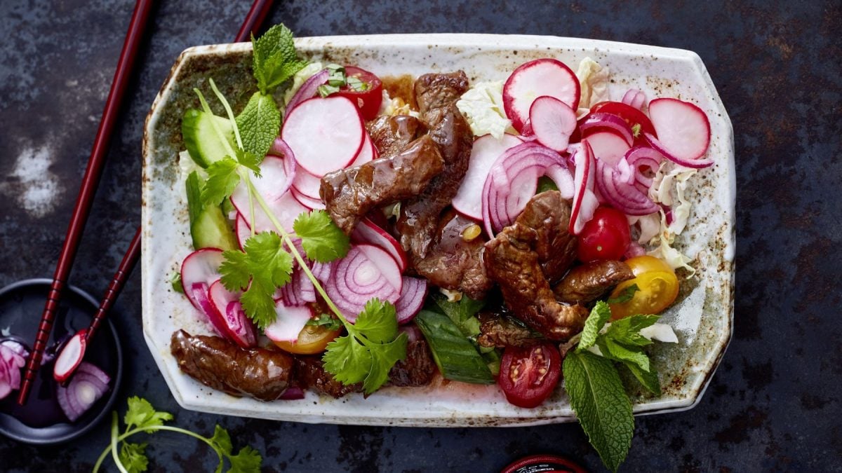 Chinakohl-Radieschen-Pickle-Slaw mit gewokten Rinderstreifen auf einem weißen Teller vor dunkelgrauem Hintergrund. Links Stäbchen und Koriander.