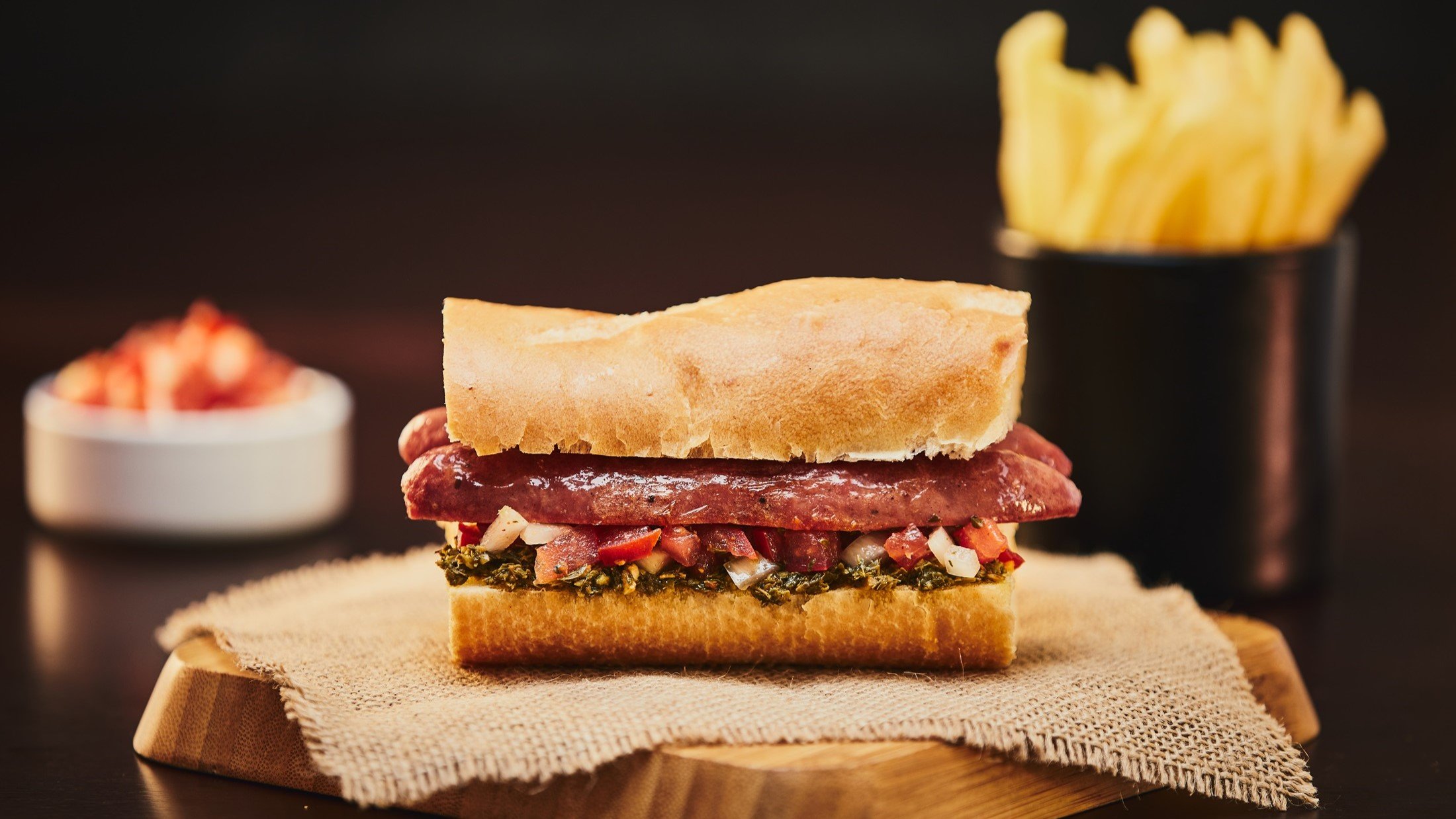 Ein Choripán, also ein Sandwich mit Chorizo und Chimichurri, auf einem Holzbrettchen und einem Tuch aus groben Leinen / Hintergrund: Pommes, eine runde Schale mit einer Sauce.