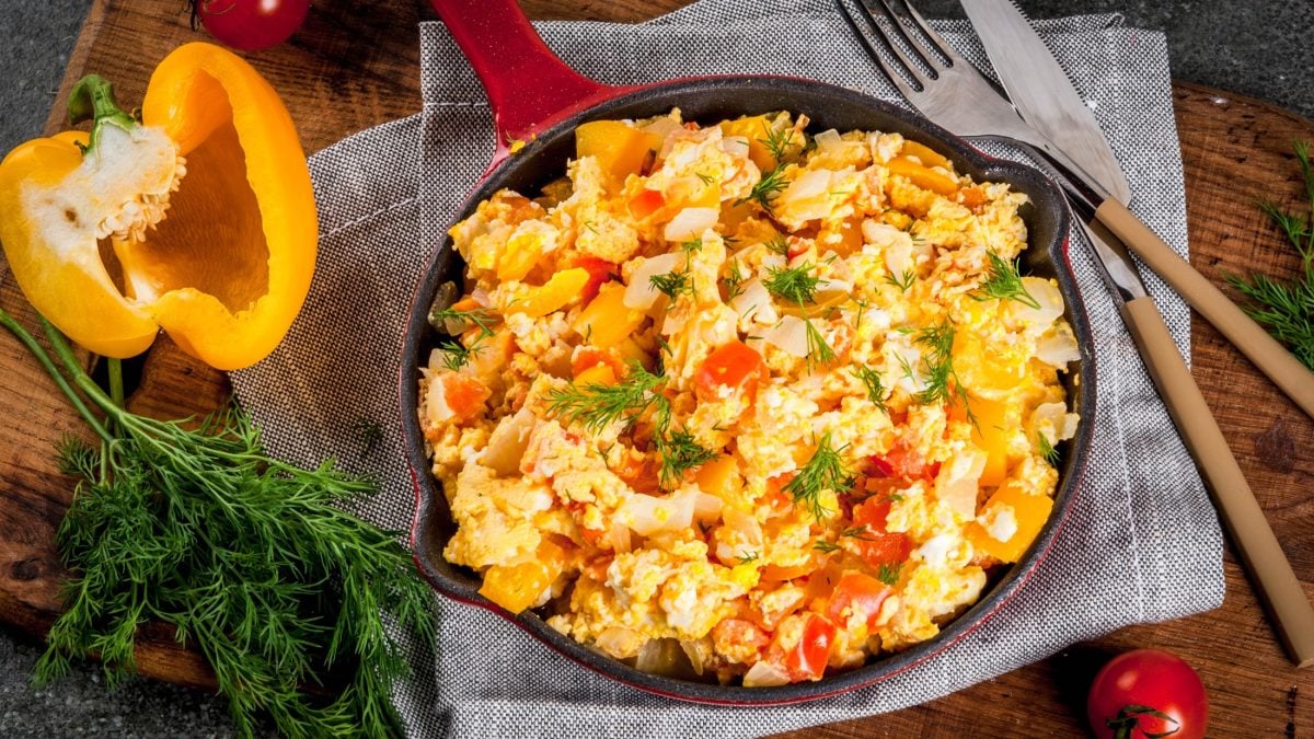 Curry-Rührei mit Paprika in einer Pfanne / halbe aufgeschnittene Paprika, Besteck / Untergrund Holz / Draufsicht schräg.