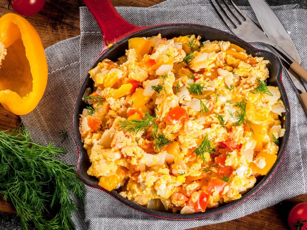 Curry-Rührei mit Paprika in einer Pfanne / halbe aufgeschnittene Paprika, Besteck / Untergrund Holz / Draufsicht schräg.