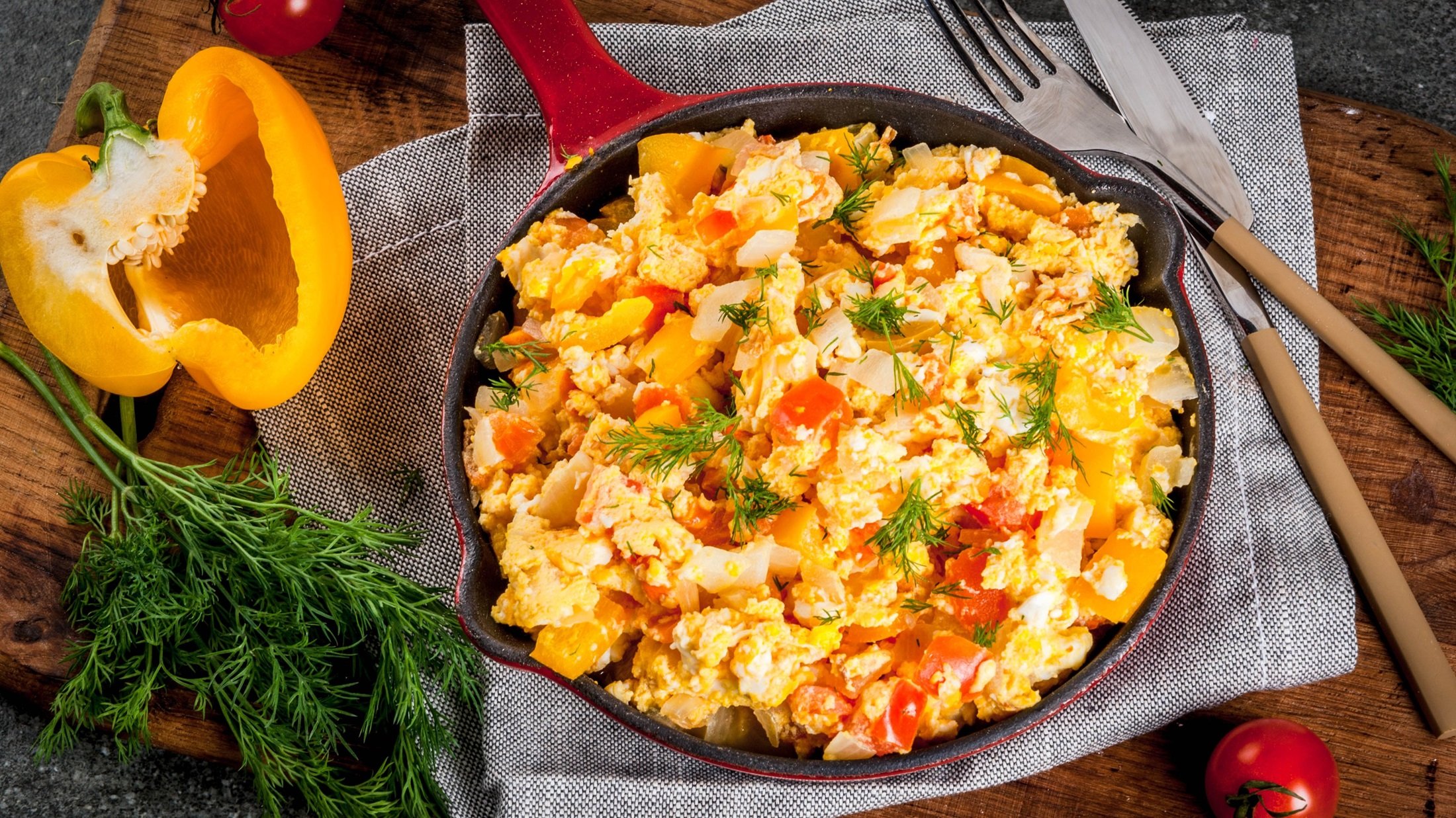 Curry-Rührei mit Paprika in einer Pfanne / halbe aufgeschnittene Paprika, Besteck / Untergrund Holz / Draufsicht schräg.