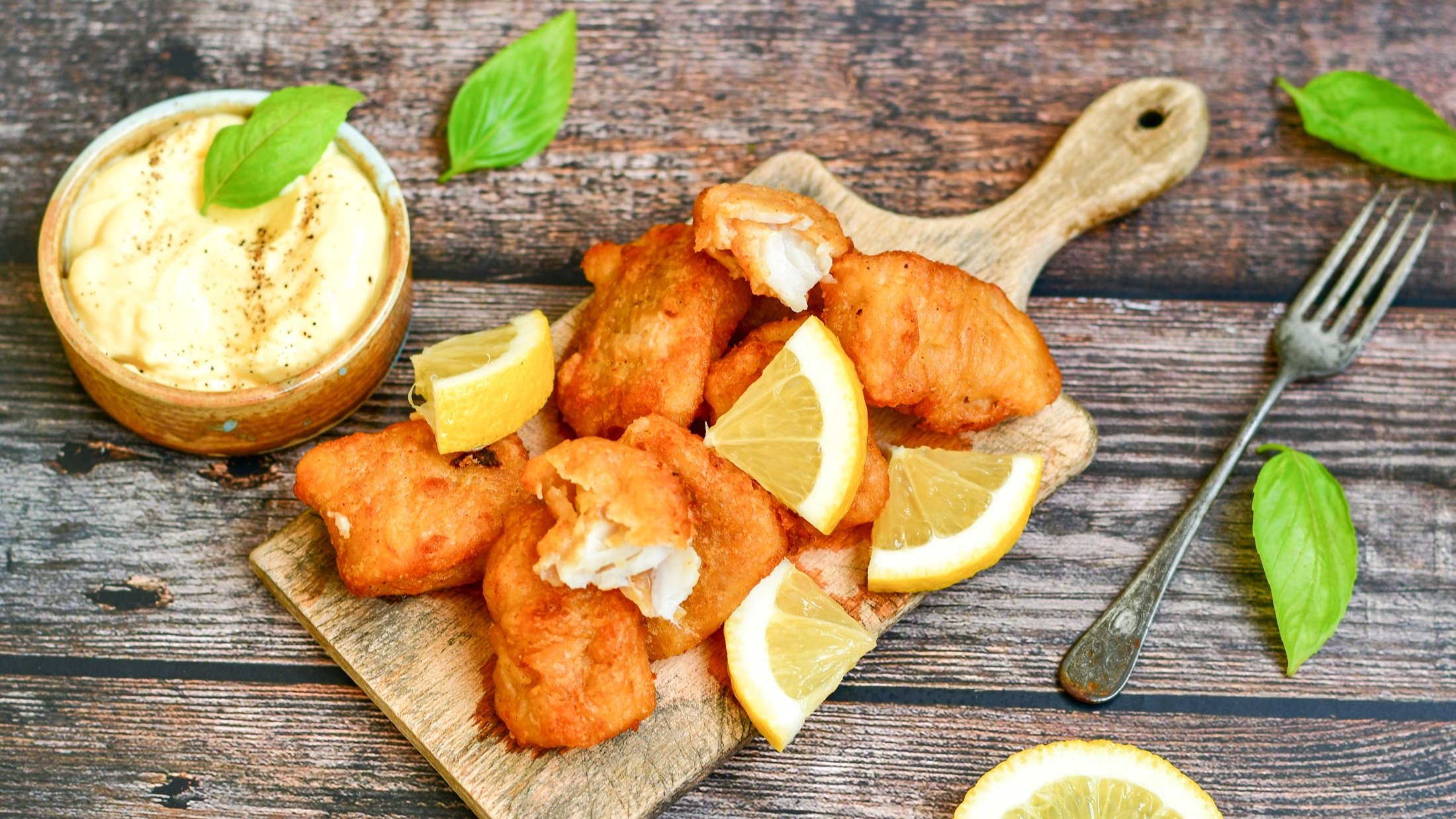 Dänische Remoulade in einer runden Schale, frittierte Fischstäbchen mit geschnittenen Zitronenscheiben auf einem Holzbrett / Draufsicht