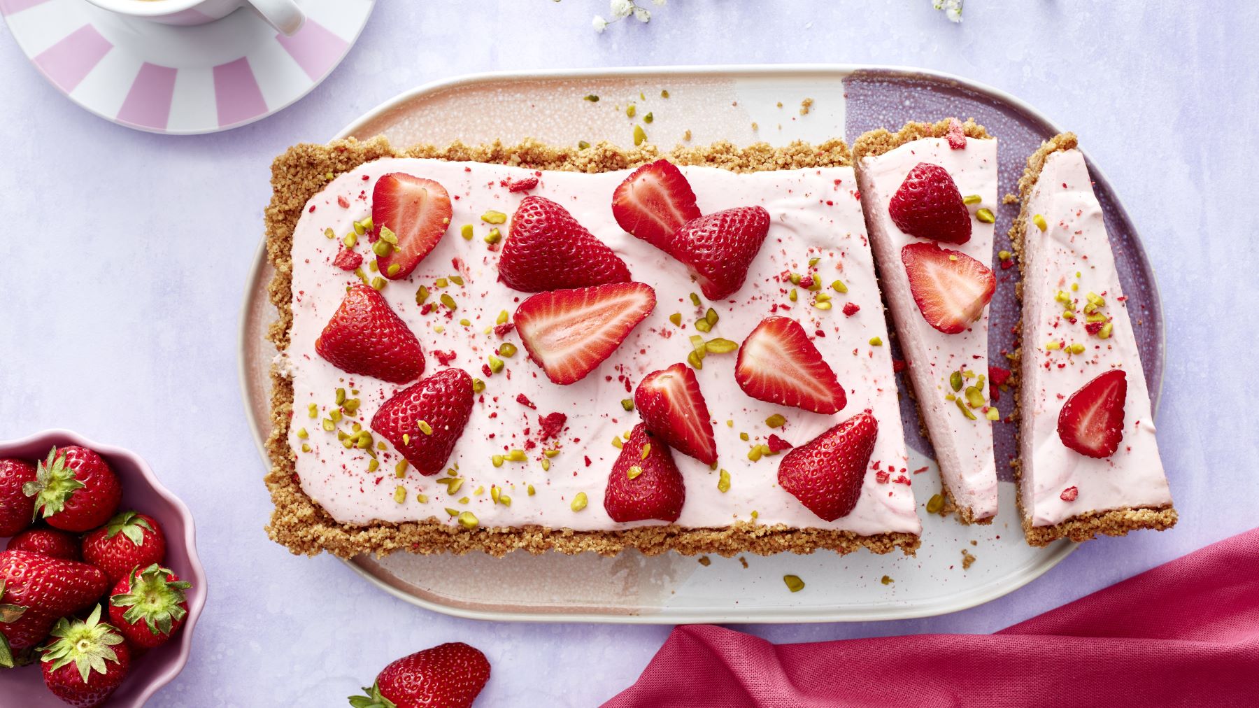 Rechteckige Erdbeer-Käsekuchen-Tarte auf einer Servierplatte, belegt mit frischen halbierten Erdbeeren und gehackten Pistazien, daneben eine Schale mit Erdbeeren und ein Stück pinker Stoff auf hellem Untergrund.