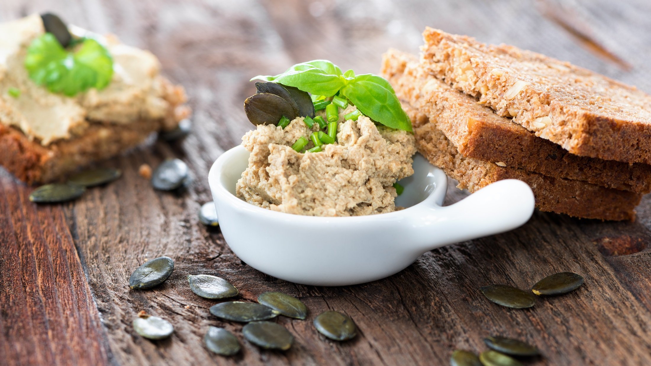 Kürbiskern-Aufstrich ist eine runden Schale. Drum herum zwei halbe Brotscheiben, lose Kürbiskerne und ein belegtes Brot mit dem Aufstrich.