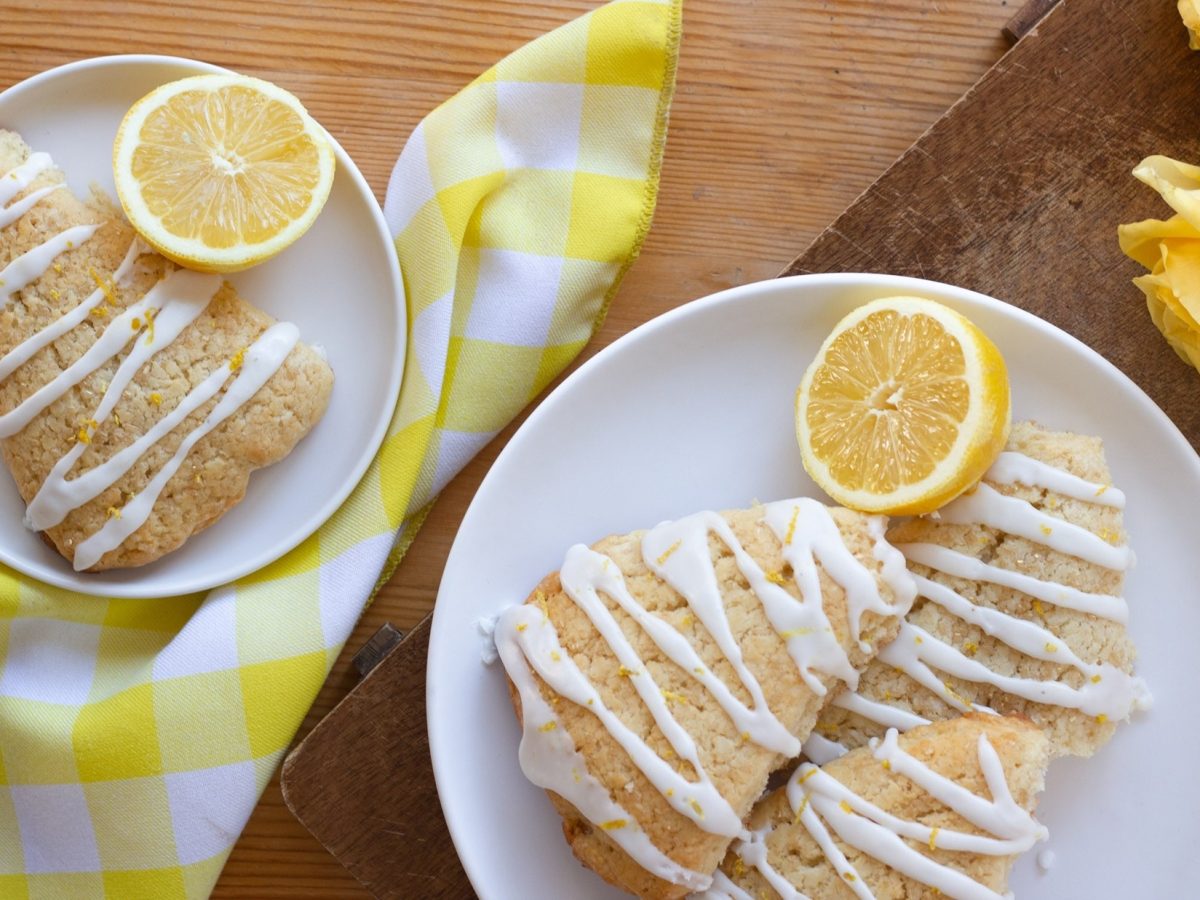Vier Lemon-Scones und Zitronenscheiben auf zwei Tellern. Daneben eine Serviette und zwei Rosenblüten. / Draufsicht