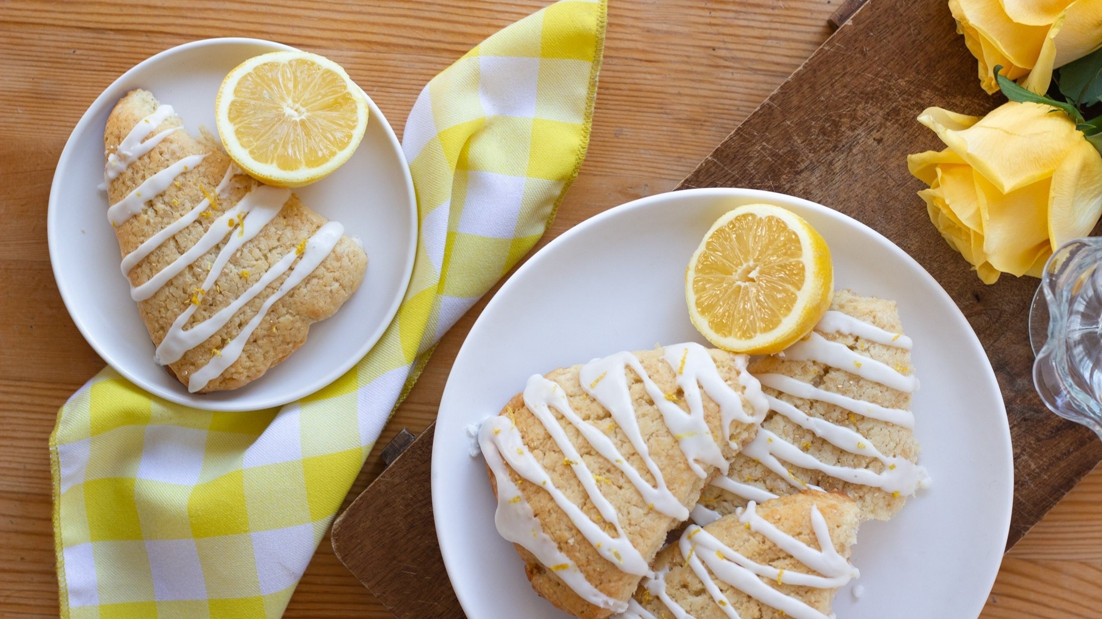 Vier Lemon-Scones und Zitronenscheiben auf zwei Tellern. Daneben eine Serviette und zwei Rosenblüten. / Draufsicht
