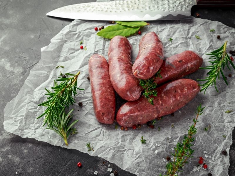 Fünf rohe Salsiccia auf Papier / frische Kräuter / ein Küchenmesser / Draufsicht, schräg
