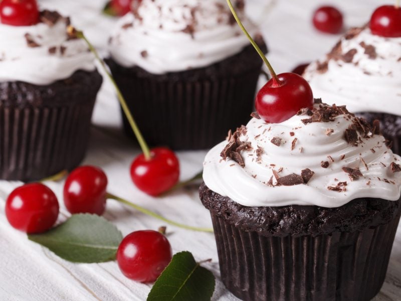 Vier Schwarzwälder Kirsch Cupcakes / heller Untergrund aus Holz / einige frische Kirschen