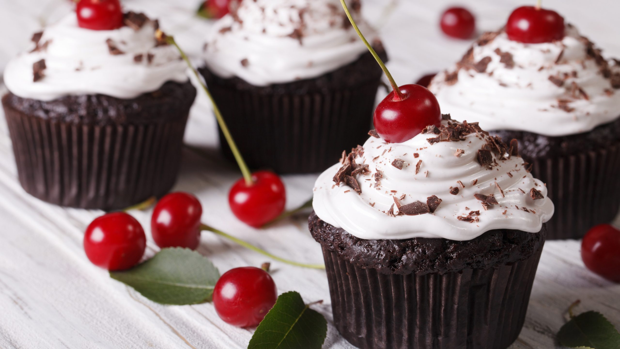 Vier Schwarzwälder Kirsch Cupcakes / heller Untergrund aus Holz / einige frische Kirschen