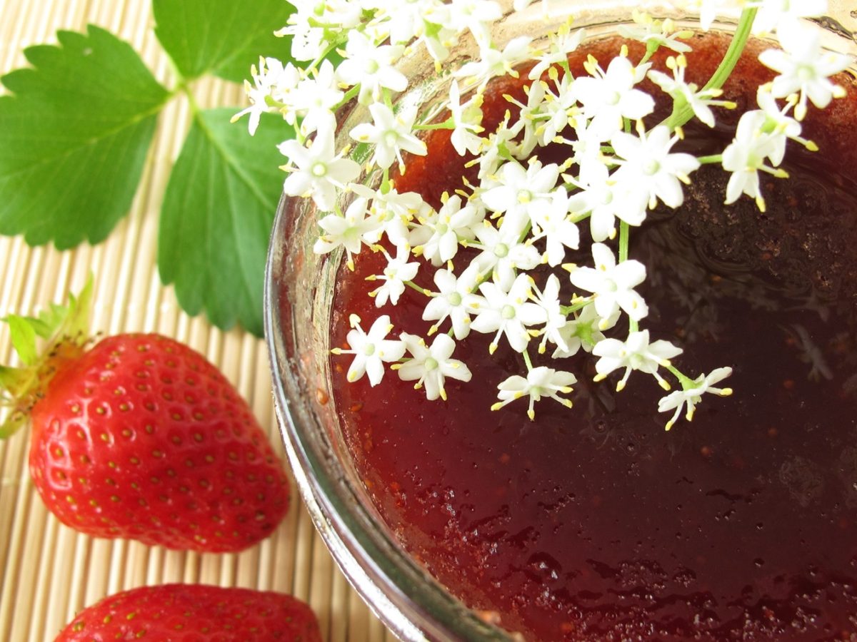 Ein Glas mit der Marmelade auf einem Holztisch, mit frischen Erdbeeren und Holunderblüten. Alles direkt von oben fotografiert.
