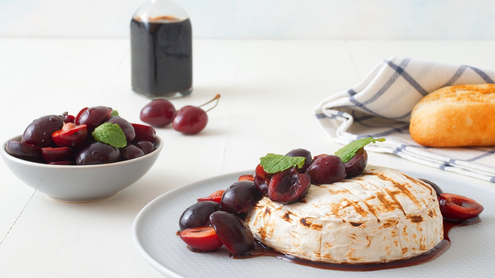 Cremiger Camembert mit Grillstreifen, garniert mit glänzenden Kirschen und Minze, daneben Brot und Schale mit Kirschkompott.