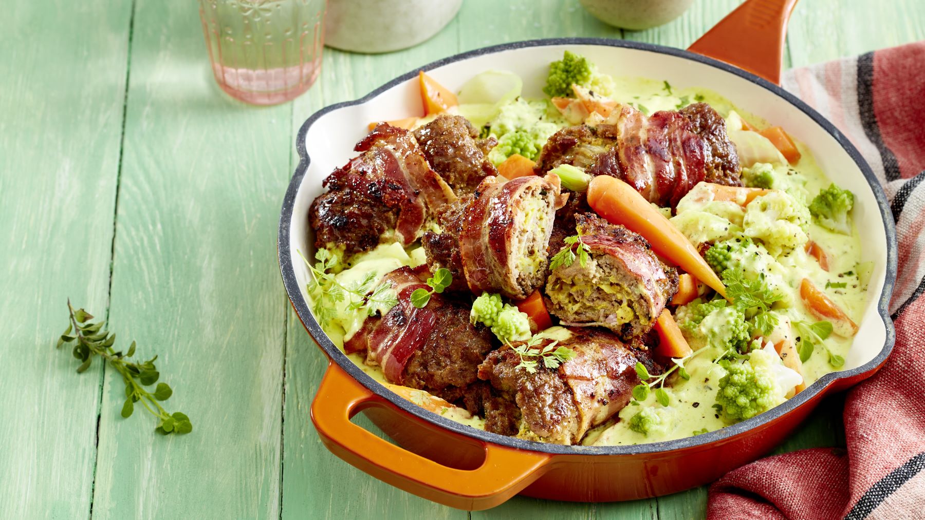 Leckere Hackröllchen-Schlemmerpfanne mit knusprigem Speck, buntem Gemüse wie Karotten und Romanesco in cremiger Sauce, serviert in einer orangefarbenen Pfanne auf grünem Holztisch.