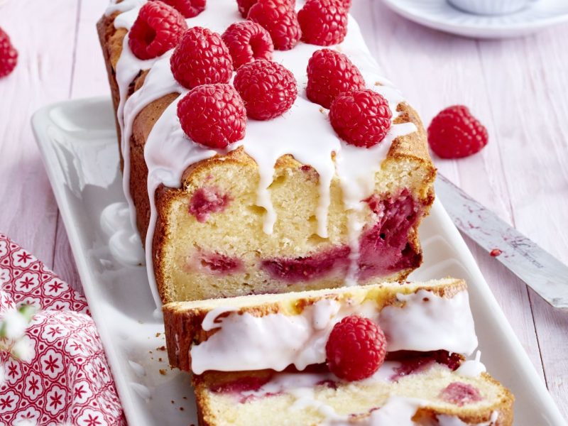 Ein saftiger Himbeer-Ricotta-Kuchen in Kastenform, mit weißem Zuckerguss überzogen und frischen Himbeeren dekoriert, angeschnitten auf einer weißen Platte, daneben ein Messer und einzelne Himbeeren auf einem hellen Holztisch.