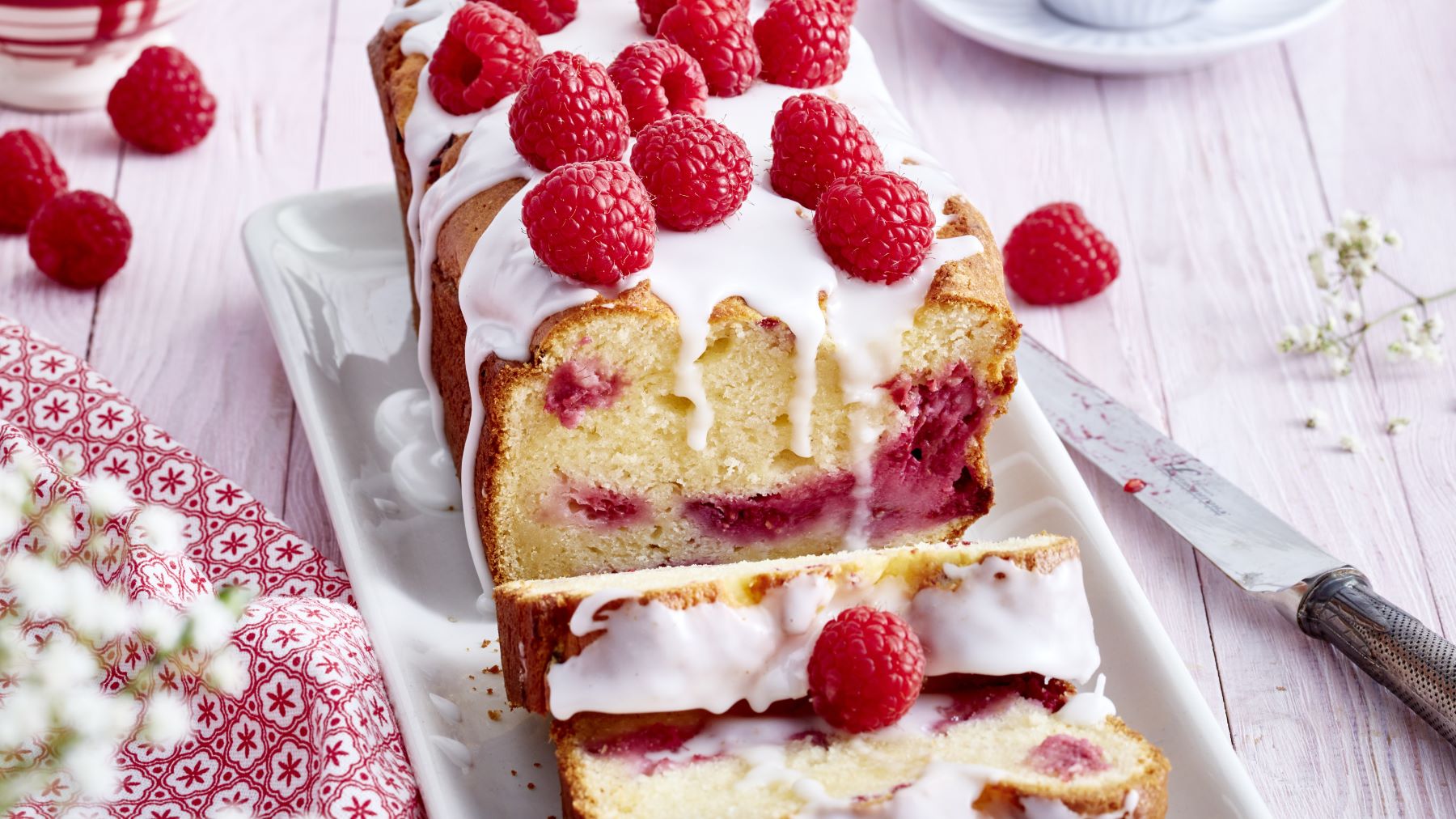 Ein saftiger Himbeer-Ricotta-Kuchen in Kastenform, mit weißem Zuckerguss überzogen und frischen Himbeeren dekoriert, angeschnitten auf einer weißen Platte, daneben ein Messer und einzelne Himbeeren auf einem hellen Holztisch.