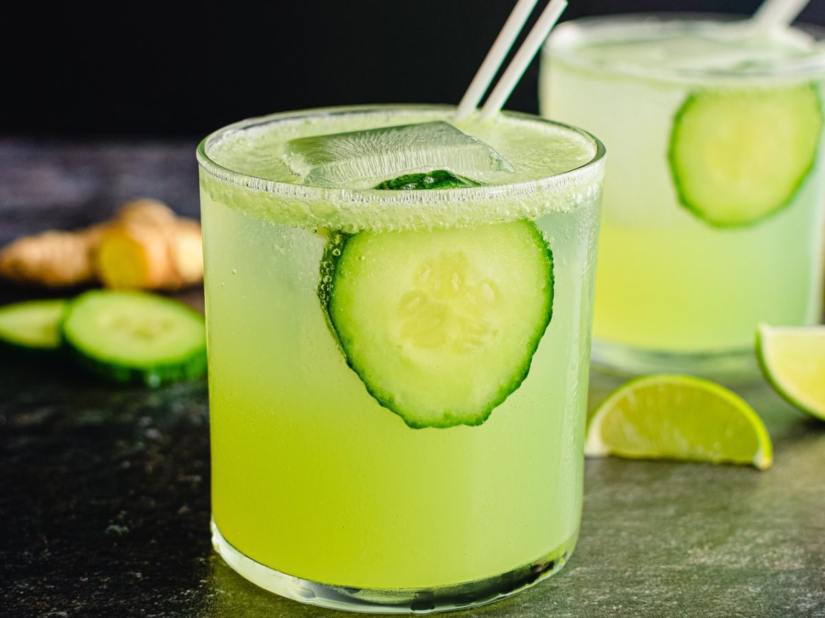 Zwei Gläser mit dem grün leuchtenden Cocktail von der Seite fotografiert. Alles auf schwarzem Untergrund mit Gurke, Limette und Ingwer.