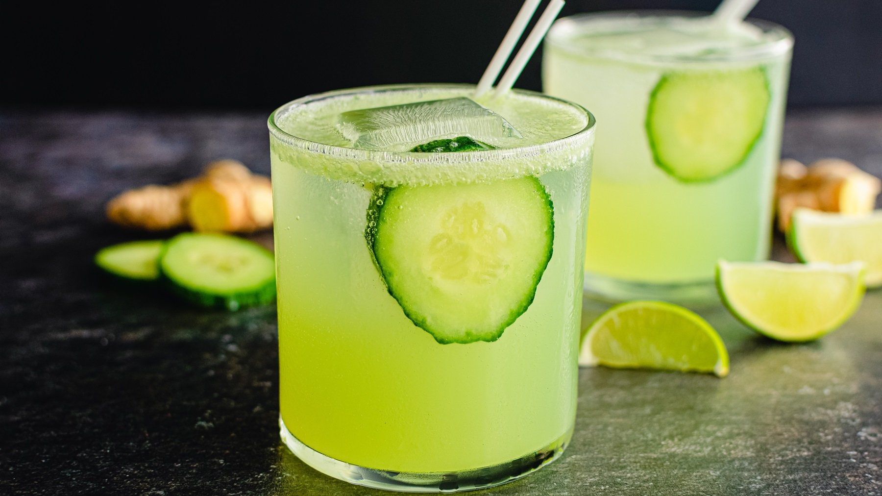Zwei Gläser mit dem grün leuchtenden Cocktail von der Seite fotografiert. Alles auf schwarzem Untergrund mit Gurke, Limette und Ingwer.