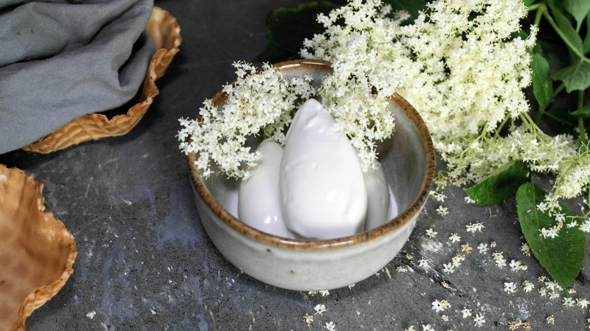 Eine Schale mit drei Eiskugeln und vielen Holunderblüten auf einem grauen Tisch von schräg oben fotografiert.