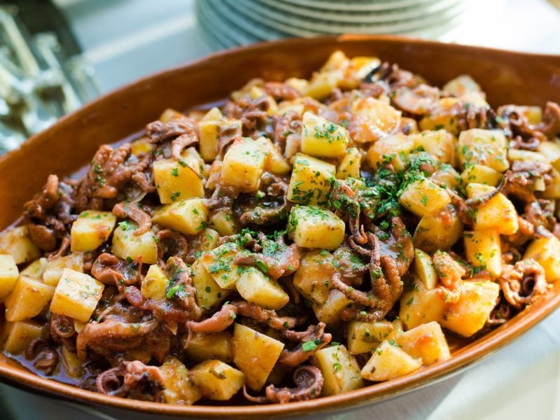 Nahaufnahme eines mediterranen Kartoffel-Oktopus-Salats mit Kräutern, serviert in rustikaler Schale auf weißer Tischdecke.