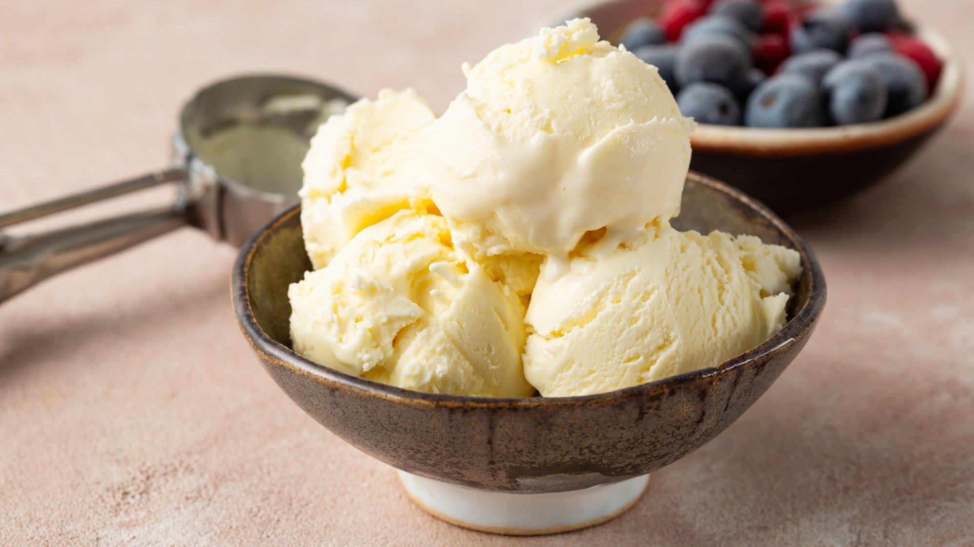 Cremige Kefir-Eiscreme in dunkler Schale auf hellem Tisch, im Hintergrund eine Schale mit Blaubeeren und Himbeeren.