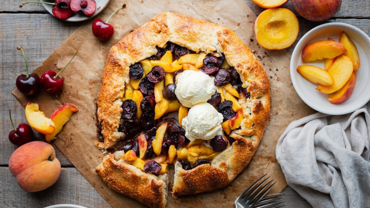 Offene Galette mit Pfirsichen und Kirschen, belegt mit zwei Kugeln Vanilleeis, auf braunem Papier und rustikalem Holztisch.