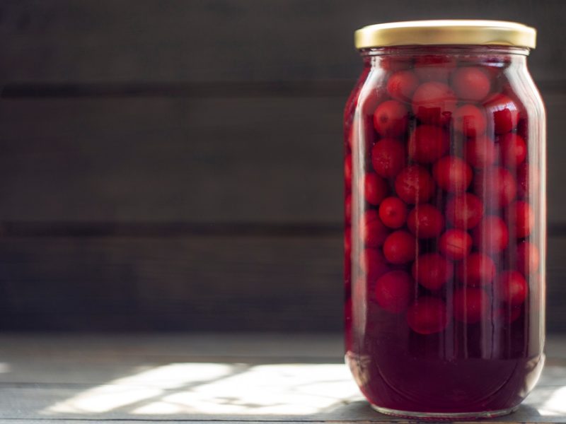 Ein hohes Einmachglas mit leuchtend roten Kirschen in dunklem Saft steht auf Holztisch vor schwarzer Wand im Sonnenlicht.