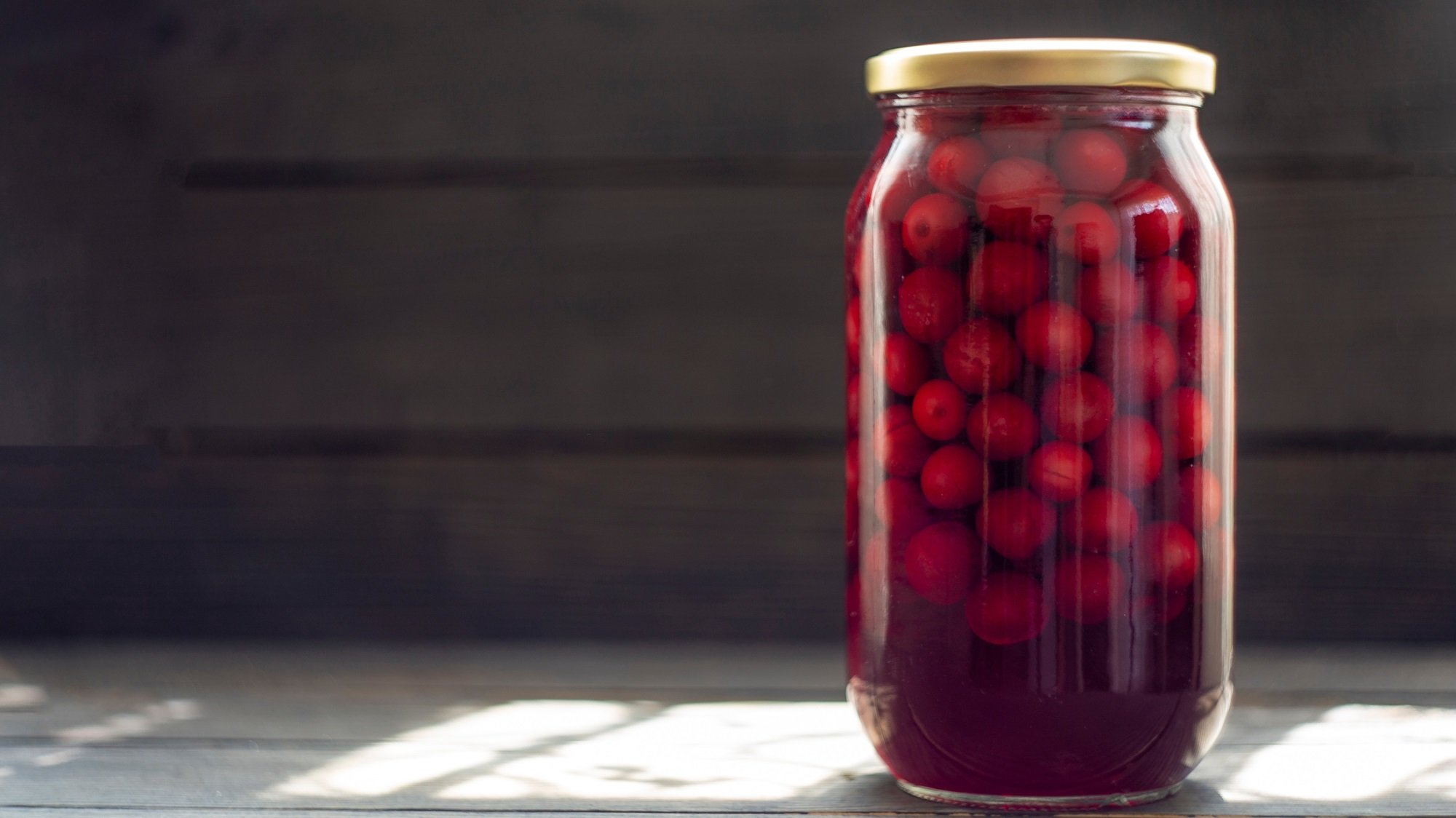 Ein hohes Einmachglas mit leuchtend roten Kirschen in dunklem Saft steht auf Holztisch vor schwarzer Wand im Sonnenlicht.