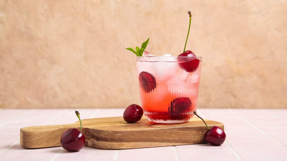Eine erfrischende Kirschlimonade mit Eiswürfeln, Minzblatt und ganzen Kirschen steht auf einem Holzbrett vor rosa Fliesen.