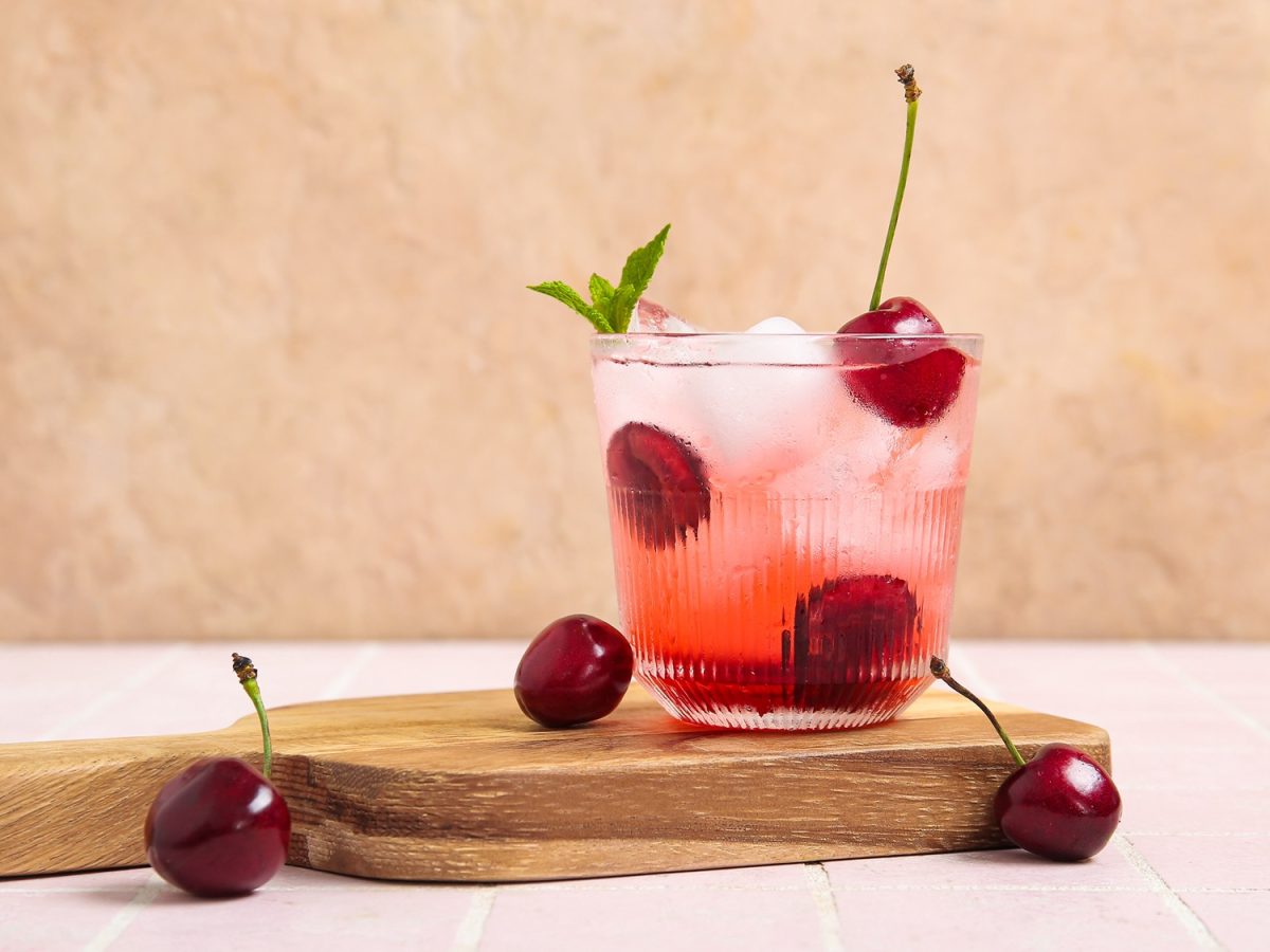 Eine erfrischende Kirschlimonade mit Eiswürfeln, Minzblatt und ganzen Kirschen steht auf einem Holzbrett vor rosa Fliesen.