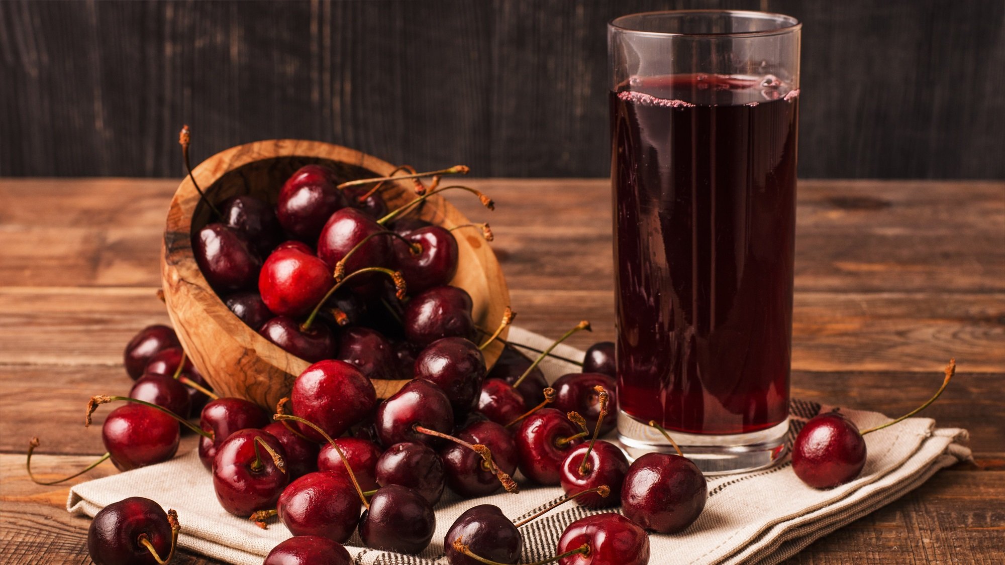 Reife, glänzende Kirschen liegen auf Leinentuch verstreut, daneben ein hohes Glas mit tiefrotem Kirschsaft.