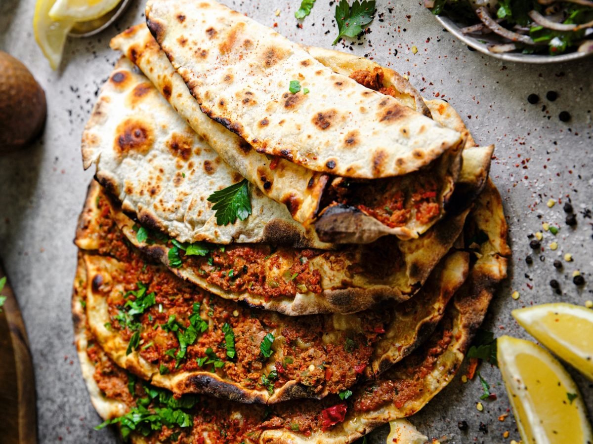 Mehrere Lahmacun auf hellgrauem Untergrund mit Zitrone und Petersilie von oben fotografiert.