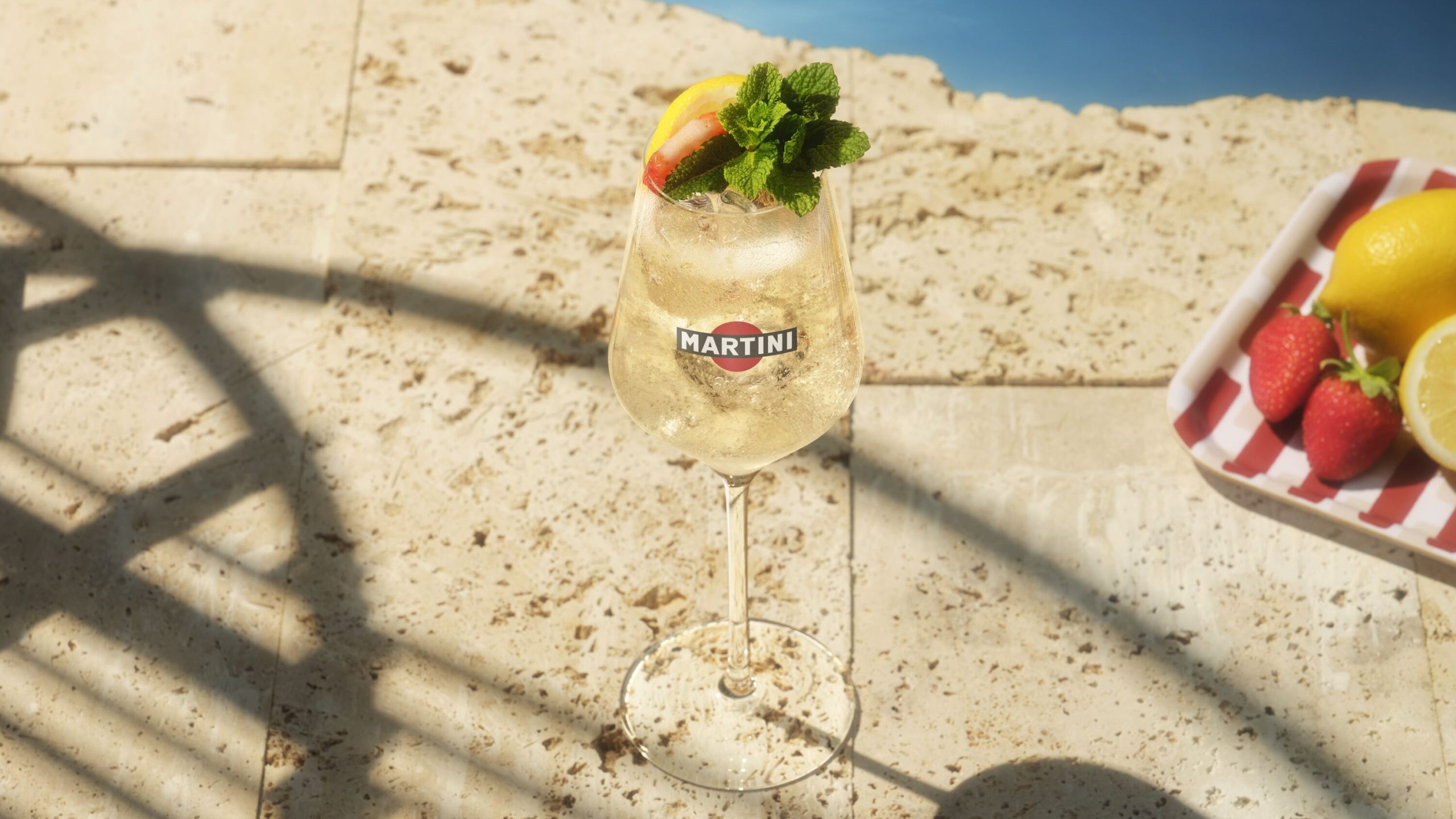 Ein Glas Martini Bianco Spritz mit Eis, Zitrone, Erdbeere und Minze steht sonnig auf hellem Stein am Poolrand.