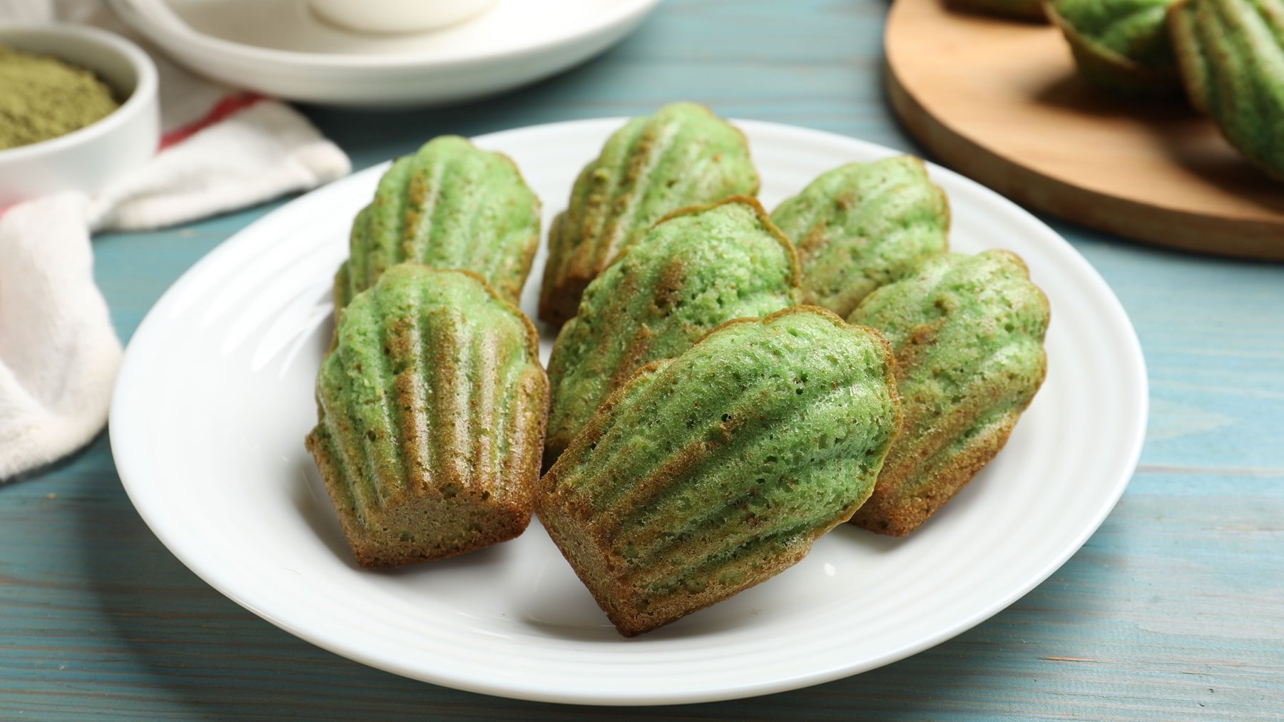 Die grünen Madeleines auf einem weißen Teller und blauem Tisch von schräg oben fotografiert.