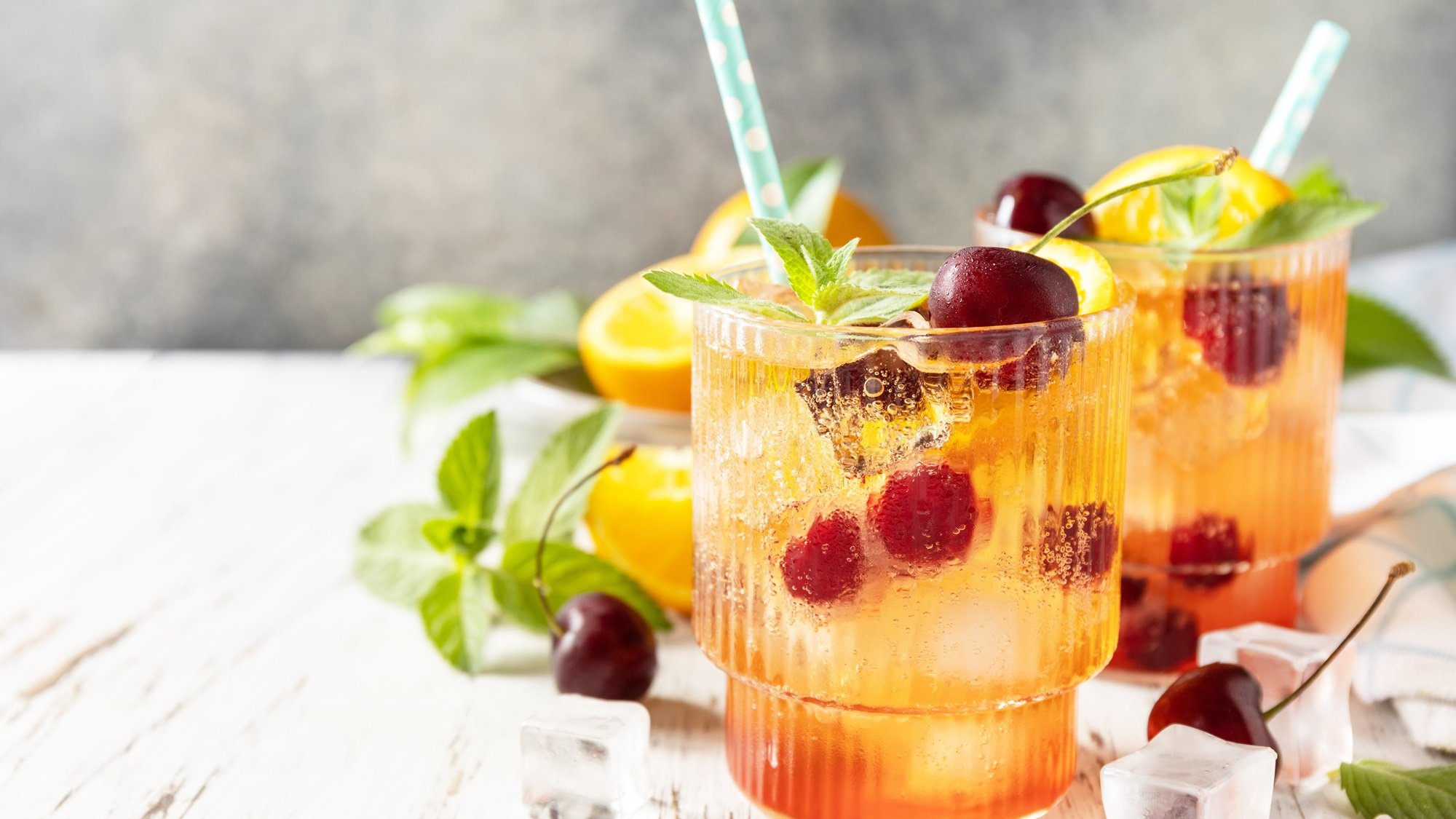 Zwei Gläser mit prickelnder Orangen-Kirsch-Limonade, garniert mit Minze, Kirschen und Eiswürfeln auf hellem Holz.