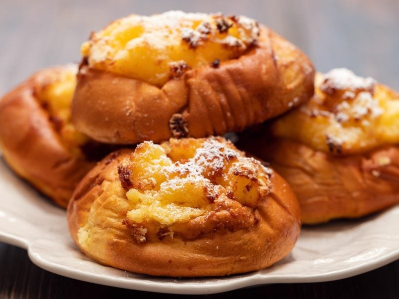 Vier goldbraune Pão de Deus mit Kokosfüllung und Puderzucker auf weißem Keramikteller vor dunklem Holzhintergrund.