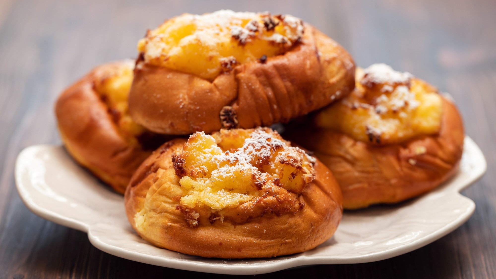 Vier goldbraune Pão de Deus mit Kokosfüllung und Puderzucker auf weißem Keramikteller vor dunklem Holzhintergrund.
